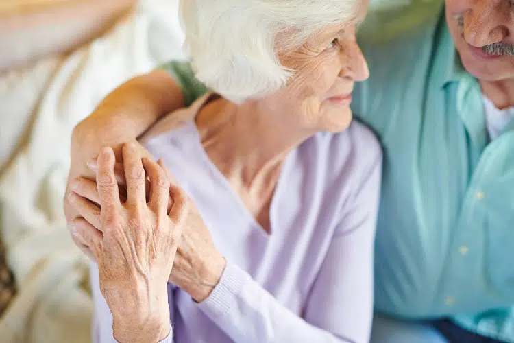 Affectionate senior woman looking at husband embracing her while enjoying free time at home in front of tv set