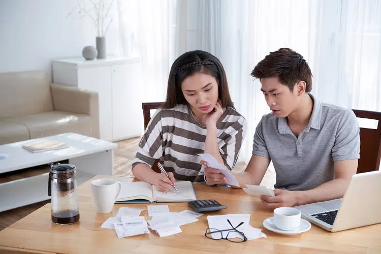 Young Asian couple gathered together at cozy living room and calculating their domestic budget, pretty woman taking necessary notes