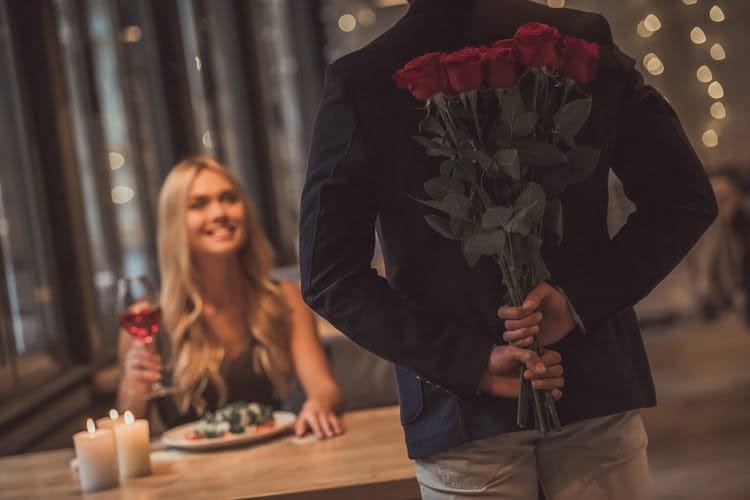 Beautiful girl is sitting at the table in restaurant and smiling while a man is holding roses behind his back