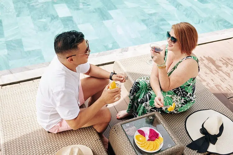 Smiling husband and wife drinking cocktails, eating fruits and talking when resting by swimming pool