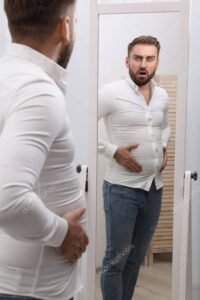 Scared man wearing tight shirt in front of mirror at home. Overweight problem