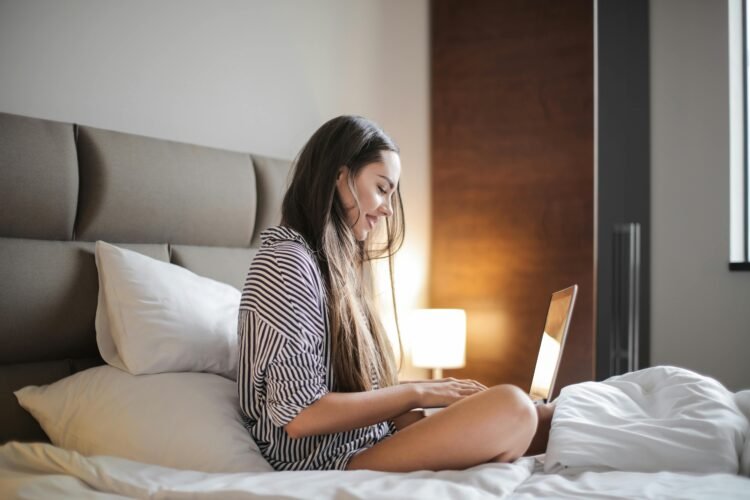 Side view photo of smiling woman in a black and white striped top sitting on a bed while using a laptop